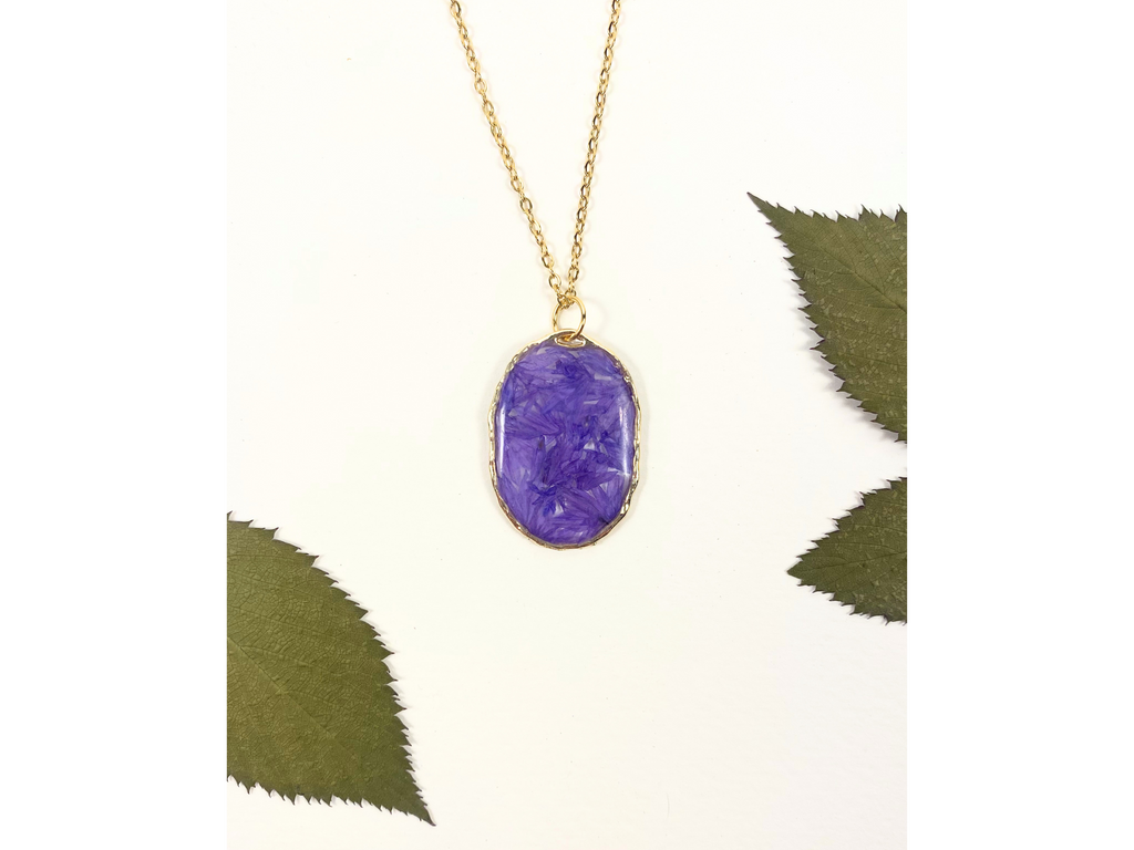 A gold chain necklace with an oval purple pendant containing dried flower inclusions, displayed on a white background with decorative green leaves.