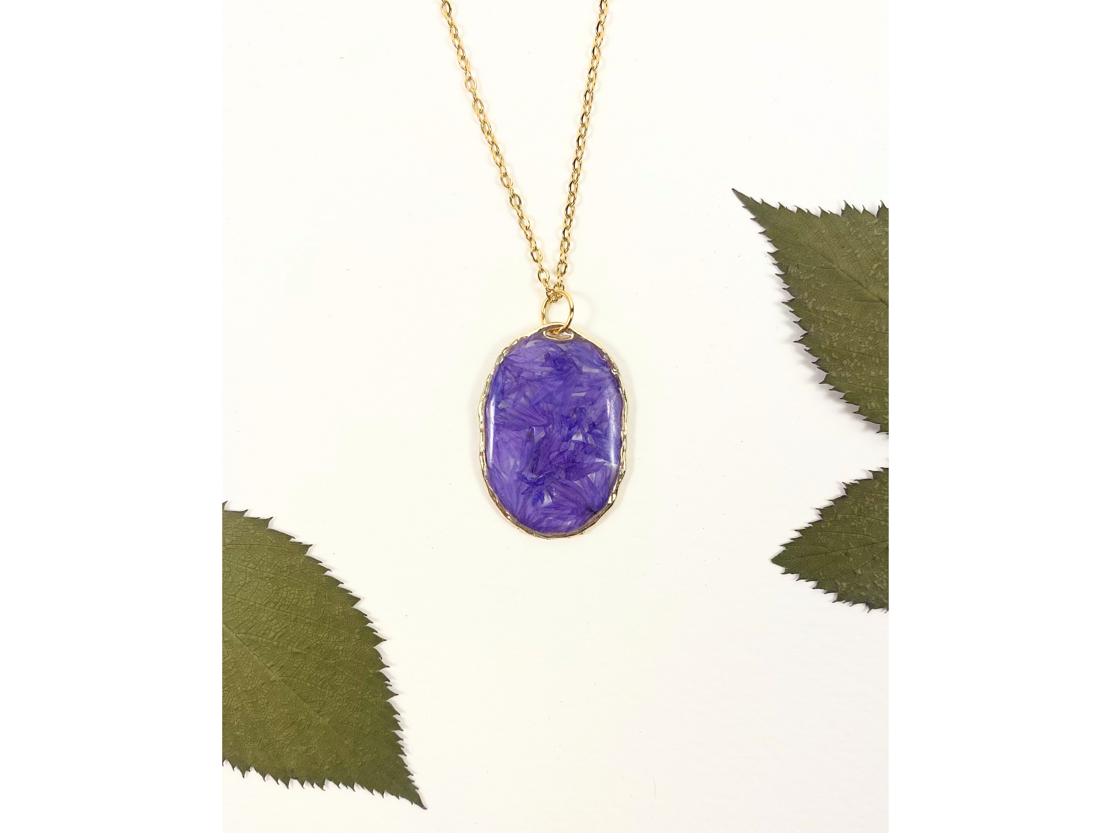 A gold chain necklace with an oval purple pendant containing dried flower inclusions, displayed on a white background with decorative green leaves.