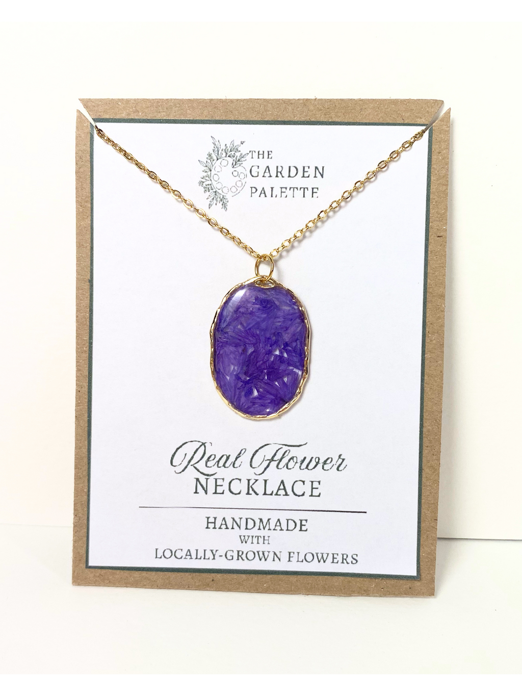 A handmade real flower necklace with purple petals, displayed on a white card with a kraft paper border. The card is branded 'The Garden Palette' and reads 'Real Flower Necklace'.