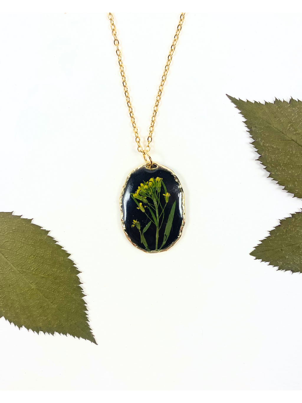 Gold chain necklace with an oval pendant featuring a black background and a pressed yellow flower, styled with green leaves on a white background.