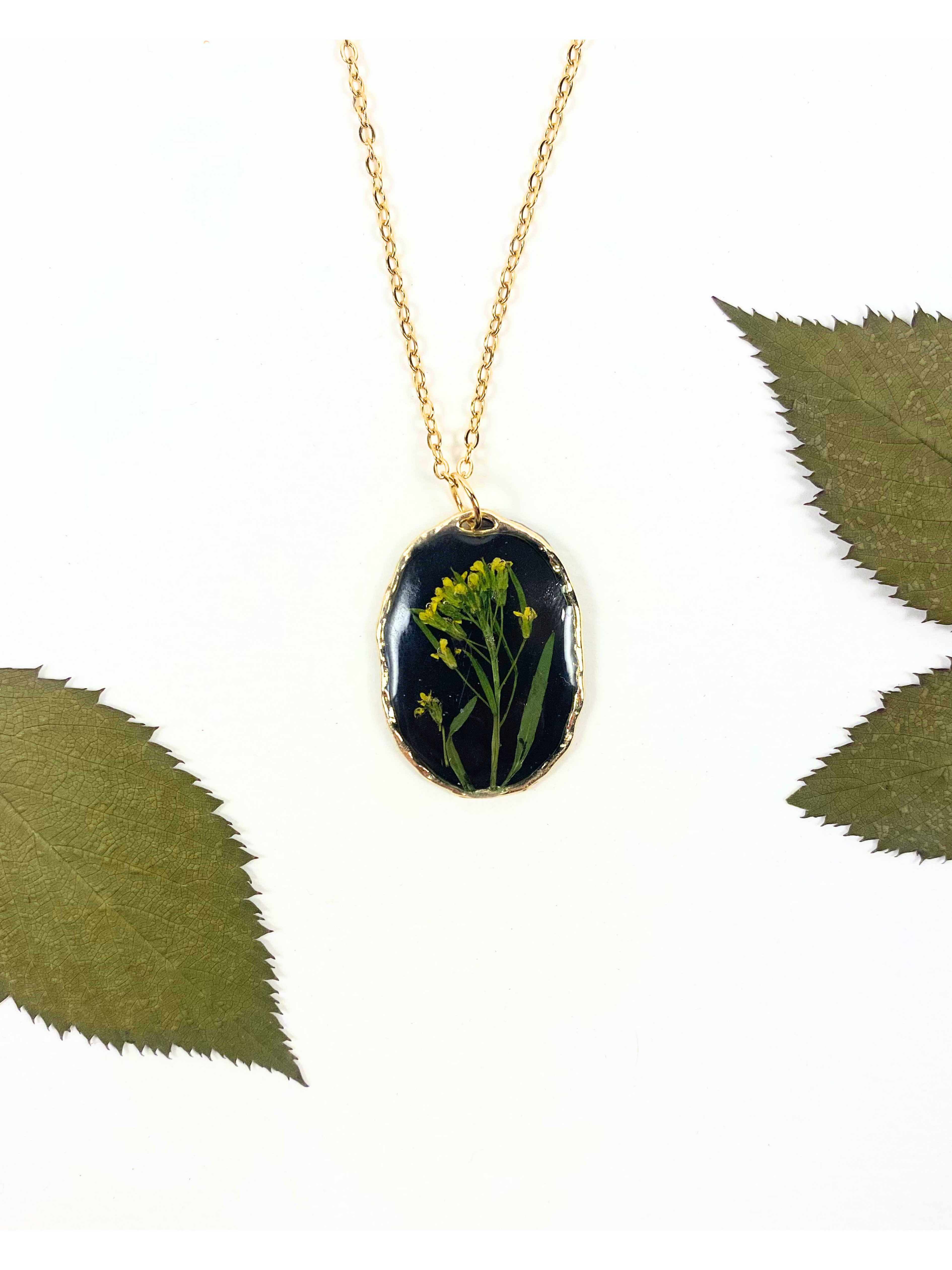 Gold chain necklace with an oval pendant featuring a black background and a pressed yellow flower, styled with green leaves on a white background.