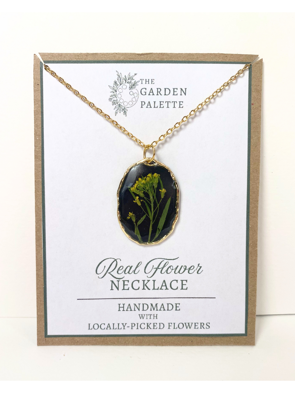 A gold-tone oval pendant necklace with a black background containing pressed yellow flowers, displayed on a white card with the brand name 'The Garden Palette' and text reading 'Real Flower Necklace'.