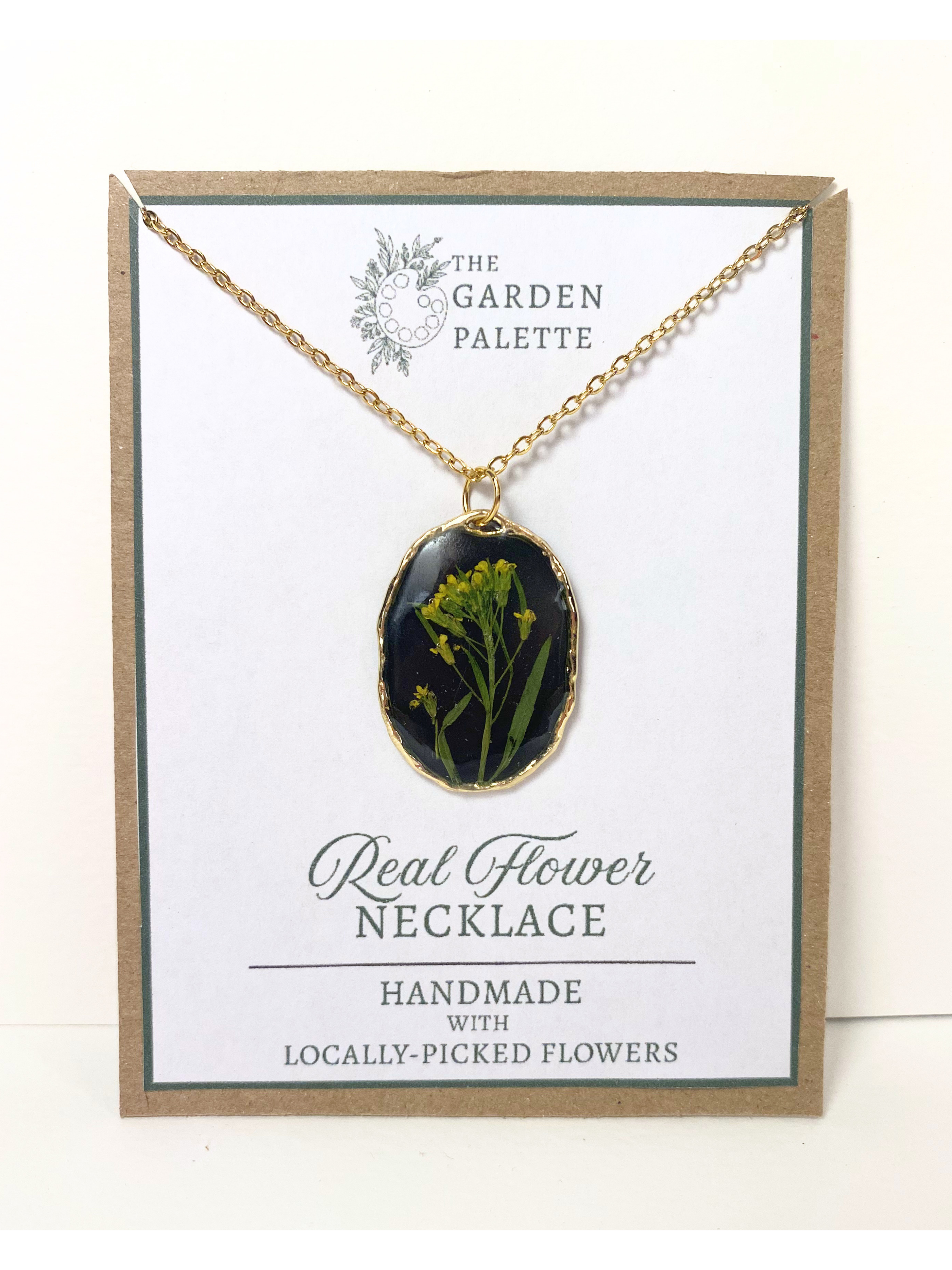 A gold-tone oval pendant necklace with a black background containing pressed yellow flowers, displayed on a white card with the brand name 'The Garden Palette' and text reading 'Real Flower Necklace'.